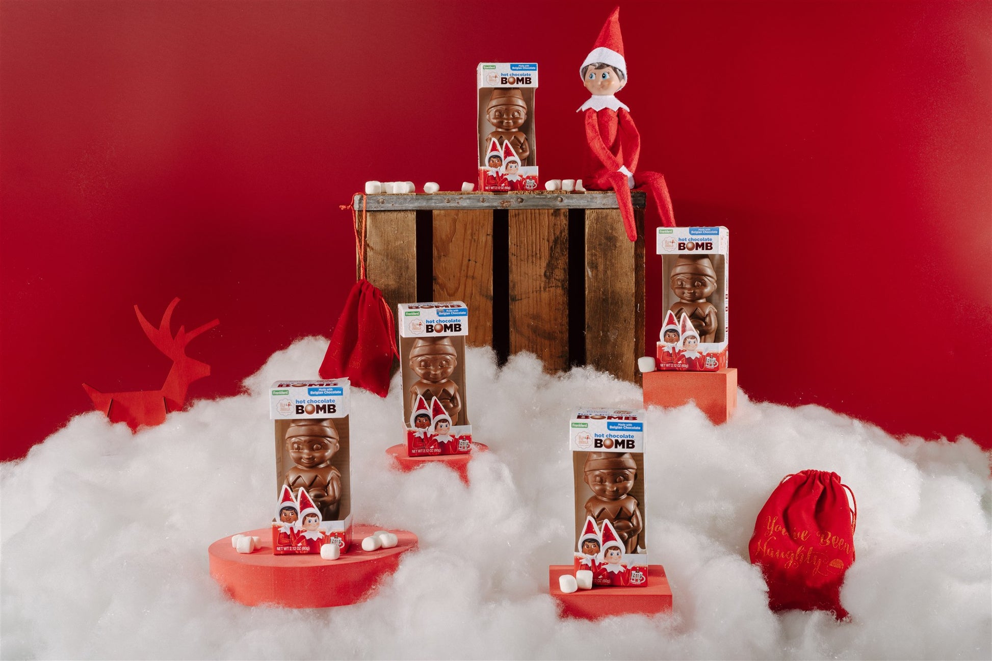 Indivdual hot chocolate bombs put on stages surrounded by fake snow with an elf on the shelf sitting on a wooden box 