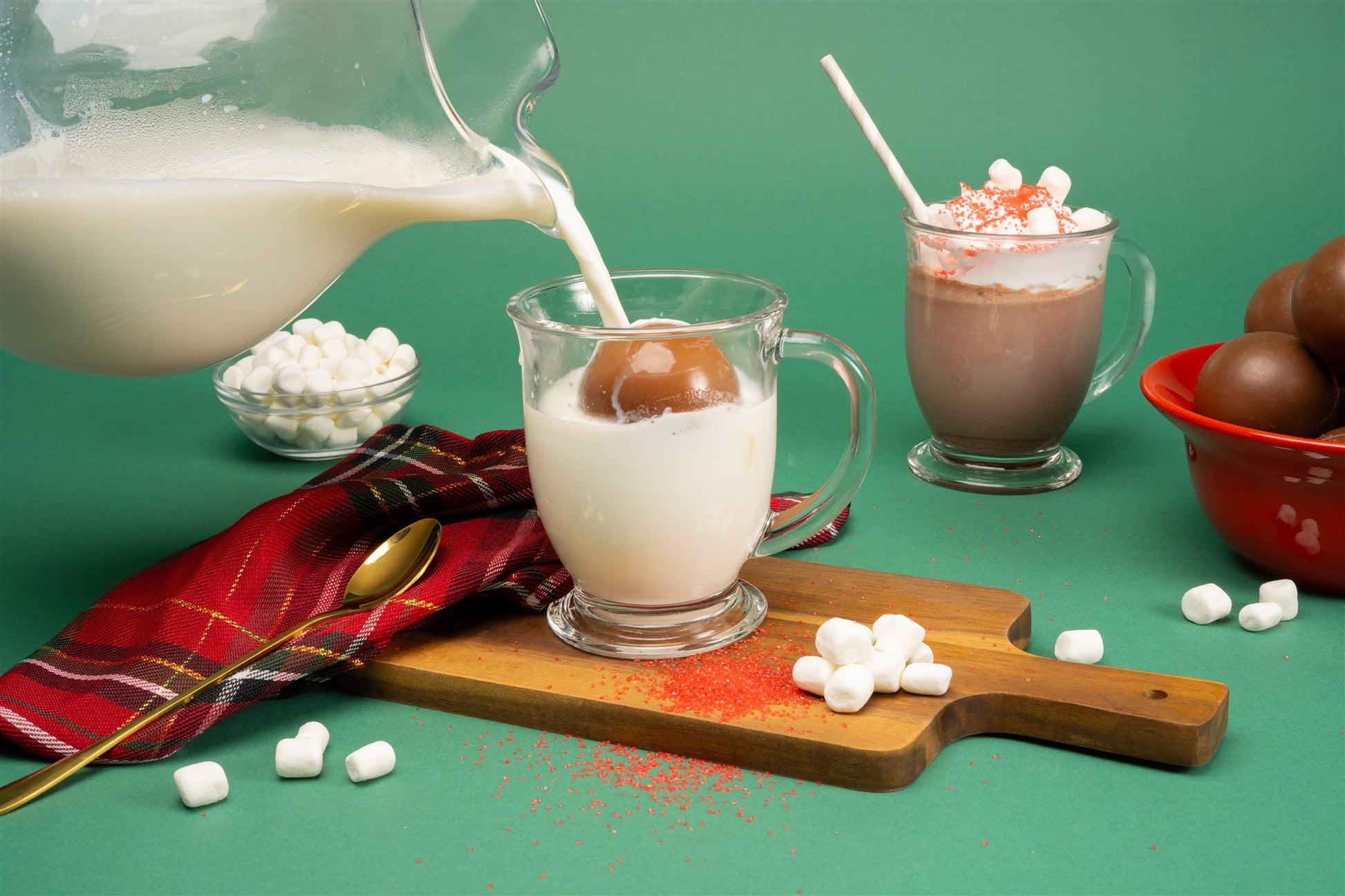 glass of milk with a hot chocolate BOMB® floating atop with a glass of milk being poured on the BOMB®