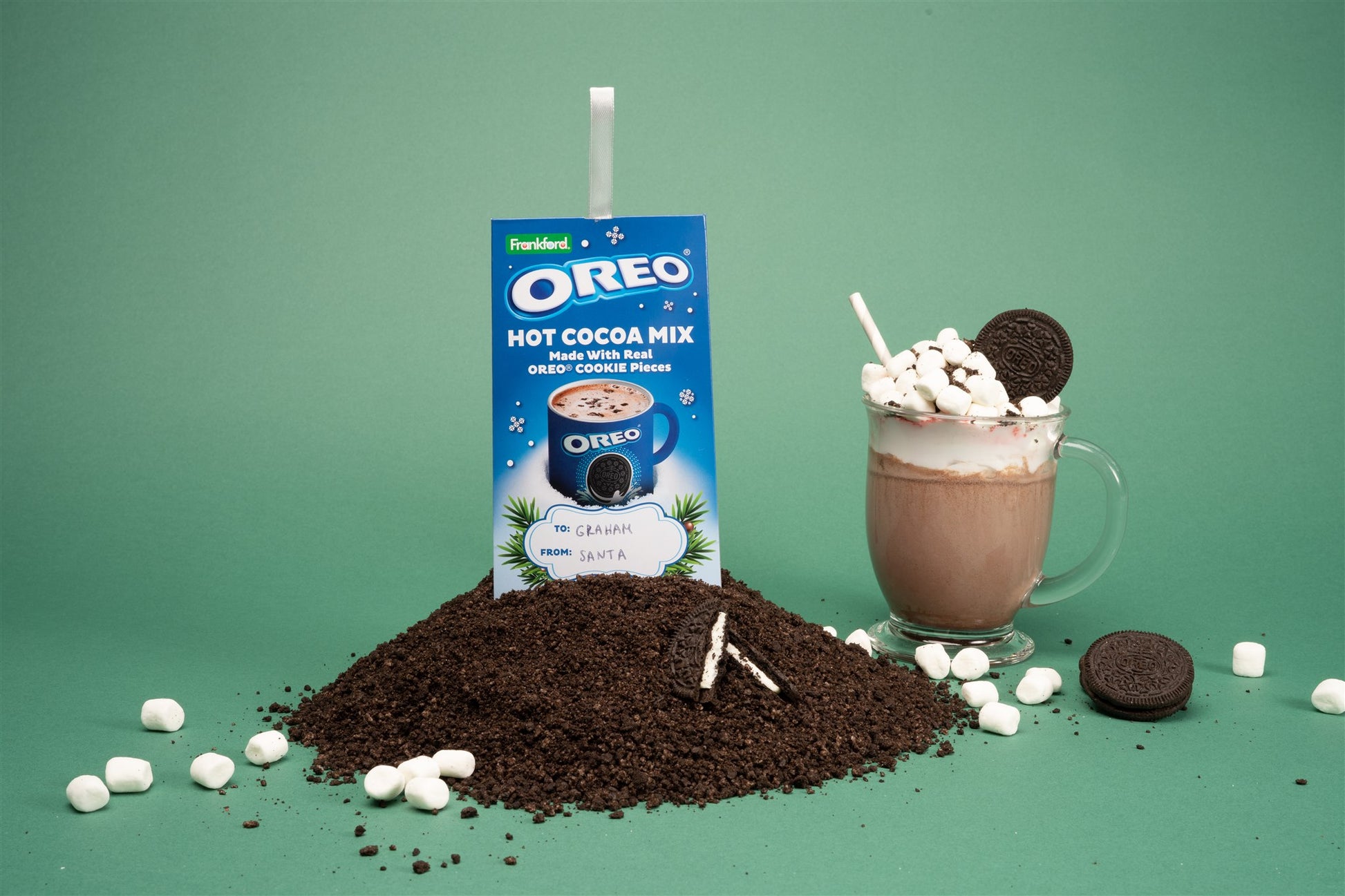 oreo hot cocoa mix packet sitting on top of crushed oreos with marshmallows scattered and a hot chocolate with oreos in the background 